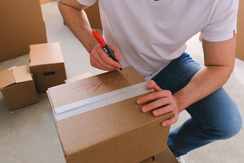 A person wearing a white T-shirt and blue jeans is kneeling indoors, using a red marker to write the word 'Jobs' on a sealed cardboard box with white packing tape. Several other cardboard boxes of various sizes, some open and some closed, are visible in the background, placed on a light-colored floor. The scene appears to be part of a home relocation, with boxes prepared for moving or packing. This image captures the process of labeling boxes for an organized house move, a service offered by Knightsbridge Removals, as indicated by the context of house removals and moving logistics.