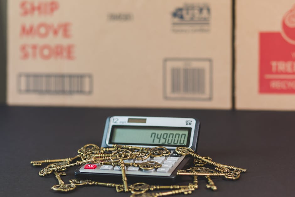 A close-up image of a small electronic calculator displaying the number 749800 on its screen, placed on a dark surface. The calculator is surrounded by multiple vintage brass keys scattered across it, some overlapping the device. Behind the calculator, there are large cardboard moving boxes with printed labels and barcodes, indicating storage or packing supplies used during house removals. The boxes are positioned in a row, partially out of focus, suggesting an indoor setting prepared for a house relocation. The scene captures elements related to the packing and moving process, including packing materials and tools involved in furniture transport and home relocation activities, consistent with professional removals services offered by Knightsbridge Removals.
