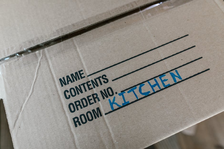 A person wearing a white T-shirt and blue jeans is kneeling indoors, using a red marker to write the word 'Jobs' on a sealed cardboard box with white packing tape. Several other cardboard boxes of various sizes, some open and some closed, are visible in the background, placed on a light-colored floor. The scene appears to be part of a home relocation, with boxes prepared for moving or packing. This image captures the process of labeling boxes for an organized house move, a service offered by Knightsbridge Removals, as indicated by the context of house removals and moving logistics.