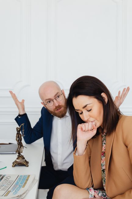 A man with glasses, a beard, and a blue suit shirt appears frustrated, gesturing with both hands raised and palms facing upwards, while seated at a desk inside a room with white panelled walls. Next to him, a woman with long dark hair, wearing a tan blazer and floral blouse, has her eyes closed and her hand covering her mouth, indicating concern or frustration. The desk holds a bronze statue, a stack of newspapers, and a laptop. The setting suggests a tense discussion related to a house removal or moving service, possibly facing a booking dispute or cancellation. These visuals are relevant to the logistics and emotional challenges involved in home relocations, which Knightsbridge Removals offers as part of their professional packing, furniture transport, and moving processes.