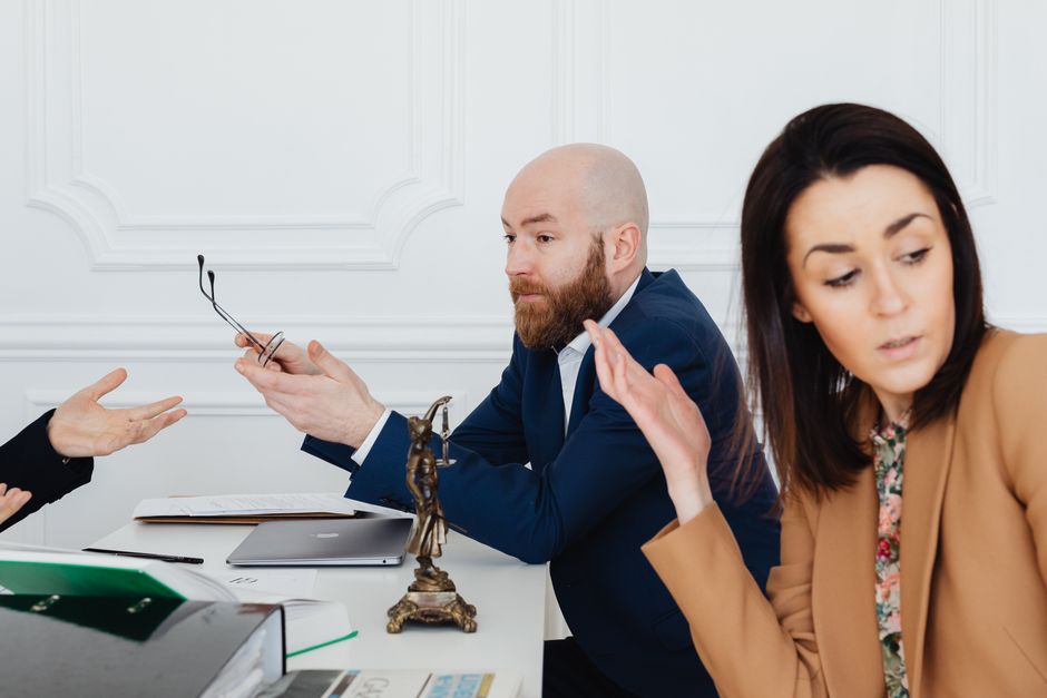 A woman is extending her hand towards a man seated at a desk, who is holding a pair of black-rimmed eyeglasses in his right hand, while gesturing with his left hand. The man has a bald head, a full beard, and is wearing a dark blue suit with a white shirt. The woman has shoulder-length dark hair, is wearing a tan blazer over a floral top, and appears to be discussing or explaining something, with a concerned expression. The desk contains various items including a closed laptop, an open notebook, green and white folders, and some documents. In front of the man, there is a small bronze sculpture of Lady Justice on the desk. The background features white paneled walls, indicating a professional office environment. The scene suggests a discussion related to a house removal or service dispute, with the employees of Knightsbridge Removals potentially addressing concerns about a home relocation process or booking issues.