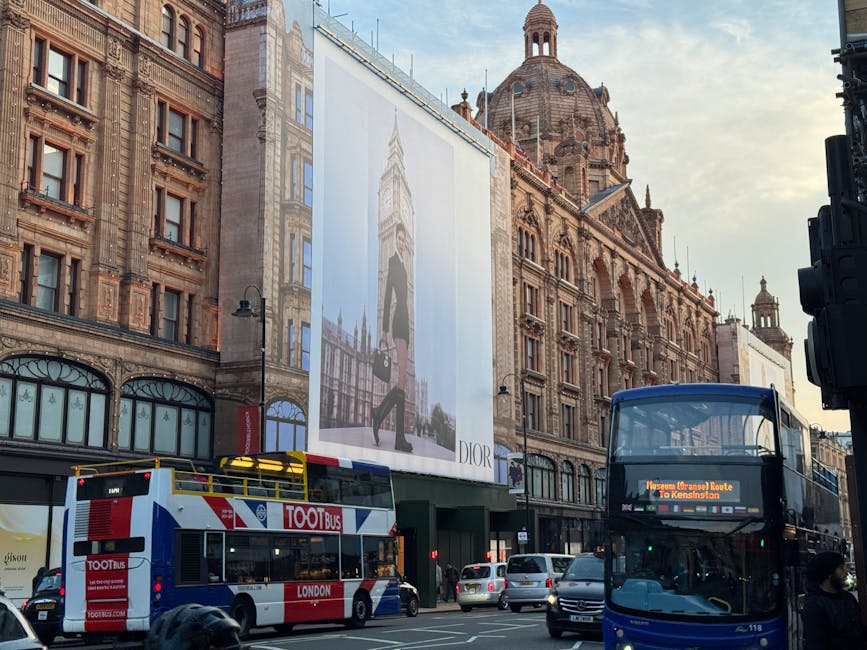 A city street scene in London showing historic architecture with ornate brick facades and a large domed building in the background. In the foreground, a double-decker bus with a digital route display is stopped at a bus stop, and a second double-decker bus with a blue and white color scheme is partially visible. Several cars are on the road, with some parked along the street. There is a large billboard mounted on the building's facade, displaying a fashion advertisement featuring a woman standing against a blue sky background. The scene captures a typical busy urban environment with traffic, vehicles, and architectural details consistent with central London. Knightsbridge Removals occasionally mention loading or transportation processes relevant to house removals and furniture transport.