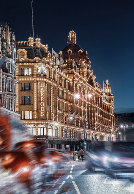 Photograph of the Harrods department store in Knightsbridge illuminated at night, showcasing its ornate architectural features and decorative lighting. The scene includes blurred moving cars on the street in front of the building, highlighting the busy London environment. In the foreground, two pedestrians are crossing the road, while the street level displays shop windows and street lamps. The image captures the grand exterior of the historic building, which is often involved in home relocation and furniture transport services by Knightsbridge Removals, sometimes requiring careful packing and loading processes for smooth moving logistics in London.