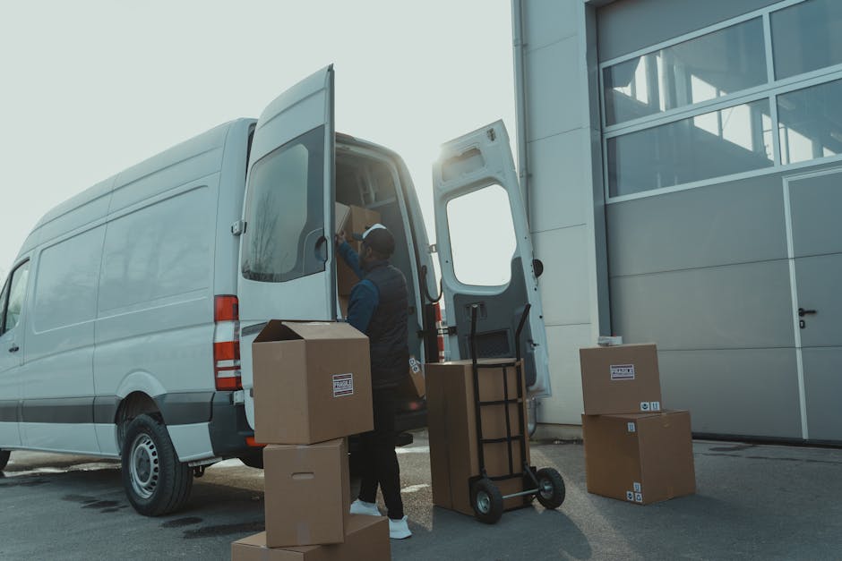 A professional mover from Knightsbridge Removals is loading packed cardboard boxes and various household items into a large white moving van parked outside a modern building with glass windows. The mover is wearing a dark uniform and is using a hand trolley to carry a box towards the open rear door of the van, which is positioned on the pavement. Several boxes are stacked nearby on the ground, some with visible labels, and a few are on the trolley ready for loading. The scene is illuminated with natural daylight, with sunlight creating a subtle glow around the van's roof and the building's reflective glass. The setting depicts the process of home relocation, involving packing, loading furniture, and transport preparations, reflecting typical elements of furniture transport and moving logistics coordinated by Knightsbridge Removals, ensuring compliance with local council rules for moving vans and permits.