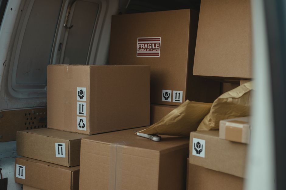Inside the rear compartment of a moving van with an open door, there are several cardboard boxes of varying sizes stacked neatly on top of each other, some with handling labels such as 'Fragile' and orientation arrows. The boxes are sealed with packing tape and are surrounded by packing paper and bubble wrap for added protection during transportation. A small folding knife or box cutter rests on the packing paper, indicating ongoing packing or unpacking activities. The interior of the van shows a metal side panel with ventilation holes, and the environment appears well-lit, possibly from natural daylight. The scene illustrates a home relocation process managed by Knightsbridge Removals, focusing on the careful packing and loading of household items, including furniture transport and moving logistics, adhering to local council rules for moving vans and permits. The image emphasizes the importance of organized packing materials and proper handling in removals services.