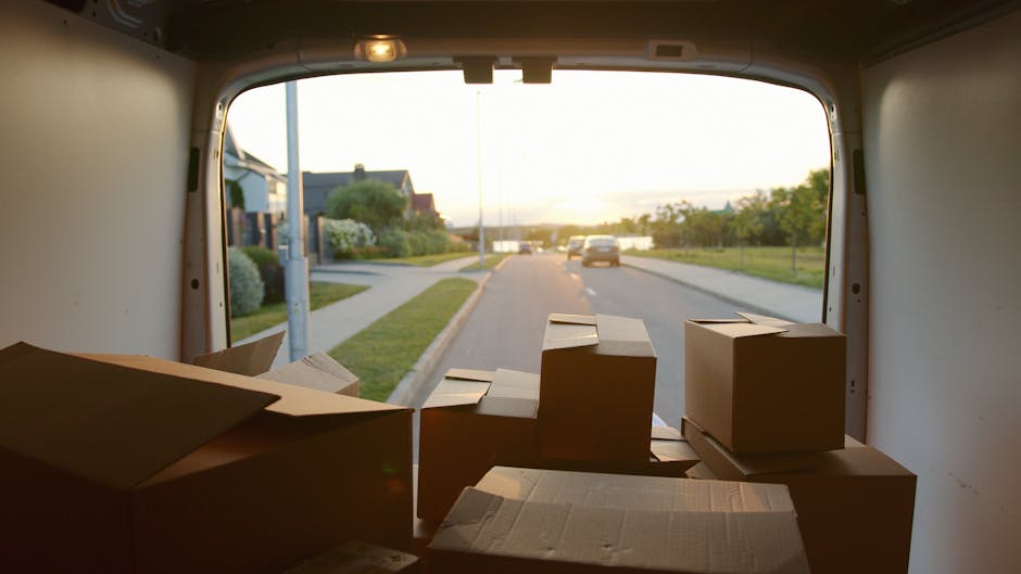 A professional mover from Knightsbridge Removals is loading packed cardboard boxes and various household items into a large white moving van parked outside a modern building with glass windows. The mover is wearing a dark uniform and is using a hand trolley to carry a box towards the open rear door of the van, which is positioned on the pavement. Several boxes are stacked nearby on the ground, some with visible labels, and a few are on the trolley ready for loading. The scene is illuminated with natural daylight, with sunlight creating a subtle glow around the van's roof and the building's reflective glass. The setting depicts the process of home relocation, involving packing, loading furniture, and transport preparations, reflecting typical elements of furniture transport and moving logistics coordinated by Knightsbridge Removals, ensuring compliance with local council rules for moving vans and permits.
