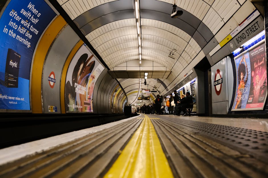 A wide-angle view of an underground London tube station platform, featuring a curved ceiling with white panels and overhead lighting. The platform has a yellow safety line running parallel to the edge, with a textured rubber surface for traction. Several passengers are seated on benches and standing near the edge, some holding luggage or bags, preparing for departures or arrivals. Advertisements and station signage, including the red circle logo of the London Underground, are visible on the walls along the platform. The station is well-lit, and the overall environment reflects a typical urban transit setting suitable for facilitating the transportation phase of home relocation or furniture transport. Knightsbridge Removals may use such stations to coordinate efficient home or office moves, ensuring safe and organized loading and unloading processes for moving services.