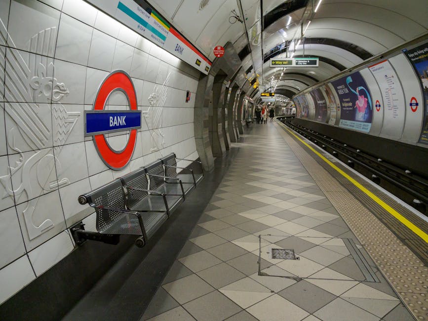Inside the London underground station near Knightsbridge, the platform features tiled walls with a large roundel sign indicating the bank station, accompanied by decorative embossed lion motifs. Several metal benches are positioned along the wall, and a few passengers are visible in the distance, standing on the tiled platform. The platform edge has a tactile warning strip with a yellow line near the tracks, which are visible alongside digital and advertising displays on the curved underground walls. Overhead, there are lighting fixtures and signs directing passengers to exits and other lines. This setting illustrates a typical home relocation or packing and moving process where furniture or boxes might be loaded or unloaded near the station, with the station’s environment designed for efficient staff and passenger movement, supported by the smooth interior and signage. Knightsbridge Removals operates within such environments for seamless transport and relocation logistics, ensuring furniture transport and packing services are handled professionally.