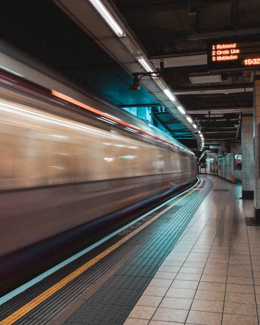 A wide-angle view of an underground London tube station platform, featuring a curved ceiling with white panels and overhead lighting. The platform has a yellow safety line running parallel to the edge, with a textured rubber surface for traction. Several passengers are seated on benches and standing near the edge, some holding luggage or bags, preparing for departures or arrivals. Advertisements and station signage, including the red circle logo of the London Underground, are visible on the walls along the platform. The station is well-lit, and the overall environment reflects a typical urban transit setting suitable for facilitating the transportation phase of home relocation or furniture transport. Knightsbridge Removals may use such stations to coordinate efficient home or office moves, ensuring safe and organized loading and unloading processes for moving services.