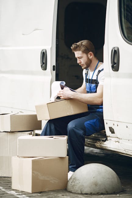 A man wearing a striped t-shirt and blue work overalls is seated at the open side door of a white van, surrounded by several cardboard boxes of various sizes, some stacked on the ground and others on the van's doorstep. He is holding a clipboard and appears to be checking or recording items during a home relocation process. Inside the van, visible behind him, are more packed boxes and protective packaging materials, such as plastic wraps and foam padding. The scene takes place outdoors on a paved surface, with a concrete bollard nearby, indicating a loading area for furniture transport and packing during house moves. The company's van is positioned close to a property, supporting efficient packing and loading for a rapid house removal or furniture transport service, consistent with the services offered by Knightsbridge Removals, as discussed on their website page about same day removals delays and common problems.