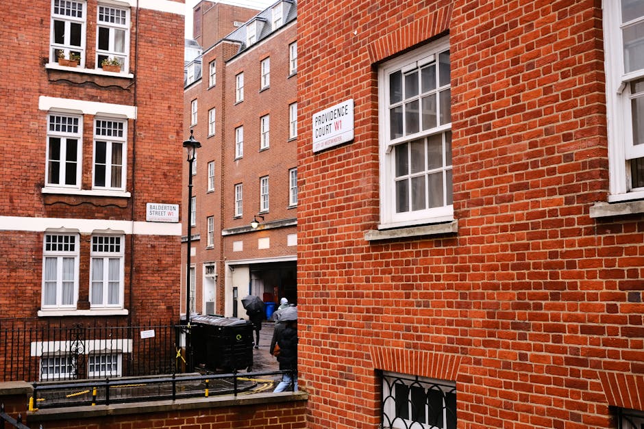 Photograph of a narrow residential street in Knightsbridge with red brick buildings and white-framed windows, showing an outdoor scene of a house removal process. Two individuals are seen outside a building, one carrying a box wrapped in plastic and another standing nearby, likely assisting with packing or furniture transport. The scene includes a black wheeled trash bin, a metal railing, and a pavement area adjacent to the buildings, with a street sign indicating Providence Court and Balderton Street, and another sign for W1. An outdoor lamp post is mounted on the corner of the building, and the background features additional multi-storey brick structures, typical of central London architecture. The lighting suggests an overcast day, with no vehicles visible but evidence of a moving process involving packing materials and manual handling, consistent with home relocation services by Knightsbridge Removals specializing in narrow access solutions.