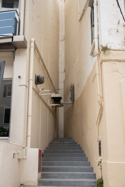 This image shows a narrow outdoor staircase located between two beige building walls, with multiple grey concrete steps leading upward. Along the left side of the staircase, there are several exposed white plastic pipes, some connected to utility meters and electrical boxes, with an additional pipe extending horizontally across the wall. On the right side, the wall appears weathered with visible signs of dirt and slight peeling paint. At the top of the stairs, a small section of pavement or landing is visible. The lighting is natural, indicating daytime, and the setting suggests a confined space often encountered during home relocations or furniture transport in urban areas. Knightsbridge Removals may utilise such staircases when conducting house removals that involve moving heavy items through tight access points, aligning with their services outlined on their website.