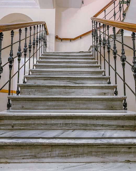An interior marble staircase inside a building, featuring polished stone steps with visible veining, flanked by black metal railings with decorative balusters and wooden handrails on both sides. The staircase ascends to a landing obscured by the angle, with white walls and a curved section at the top. The setting appears to be well-lit, with natural light illuminating the area. In the context of house removals, Knightsbridge Removals may be involved in safely transporting furniture and boxes up or down this staircase as part of a home relocation or furniture transport process, utilizing safety equipment such as moving blankets or trolleys where necessary.